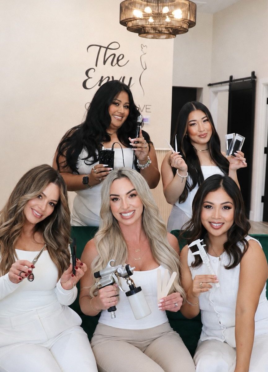 Five smiling beauty artists dressed in white pose together inside The Envy Collective, holding the tools of their trade. Nicole Velazco of Bronze Envy is front and center with an airbrush spray tan gun, and The Envy Collective logo is on the wall behind them.